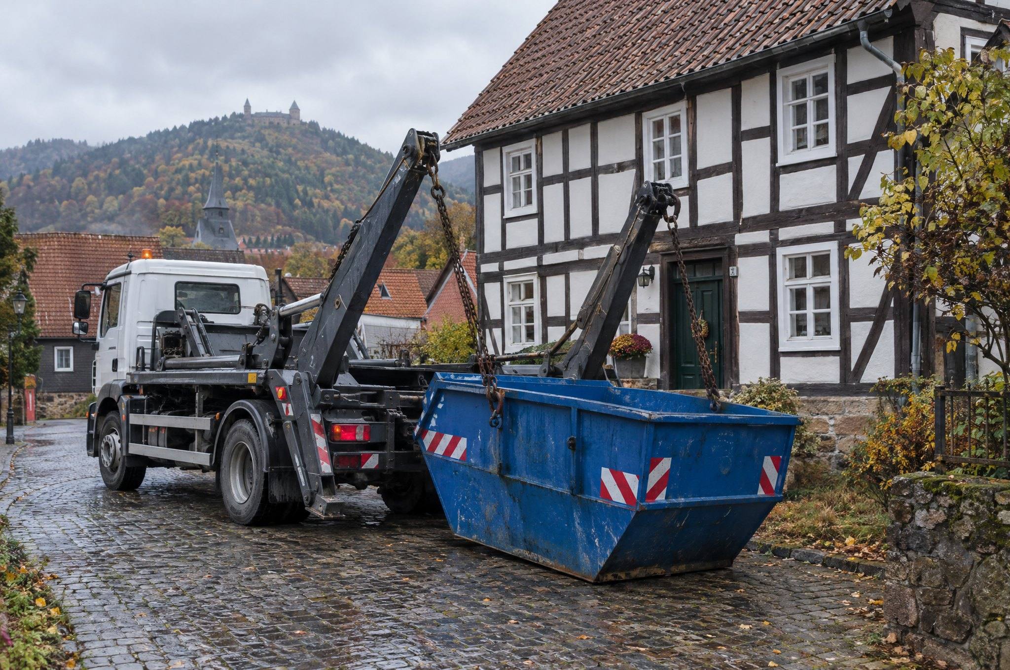 Absetzkipper stellt einen blauen Container auf einer gepflasterten Einfahrt vor einem Fachwerkhaus ab