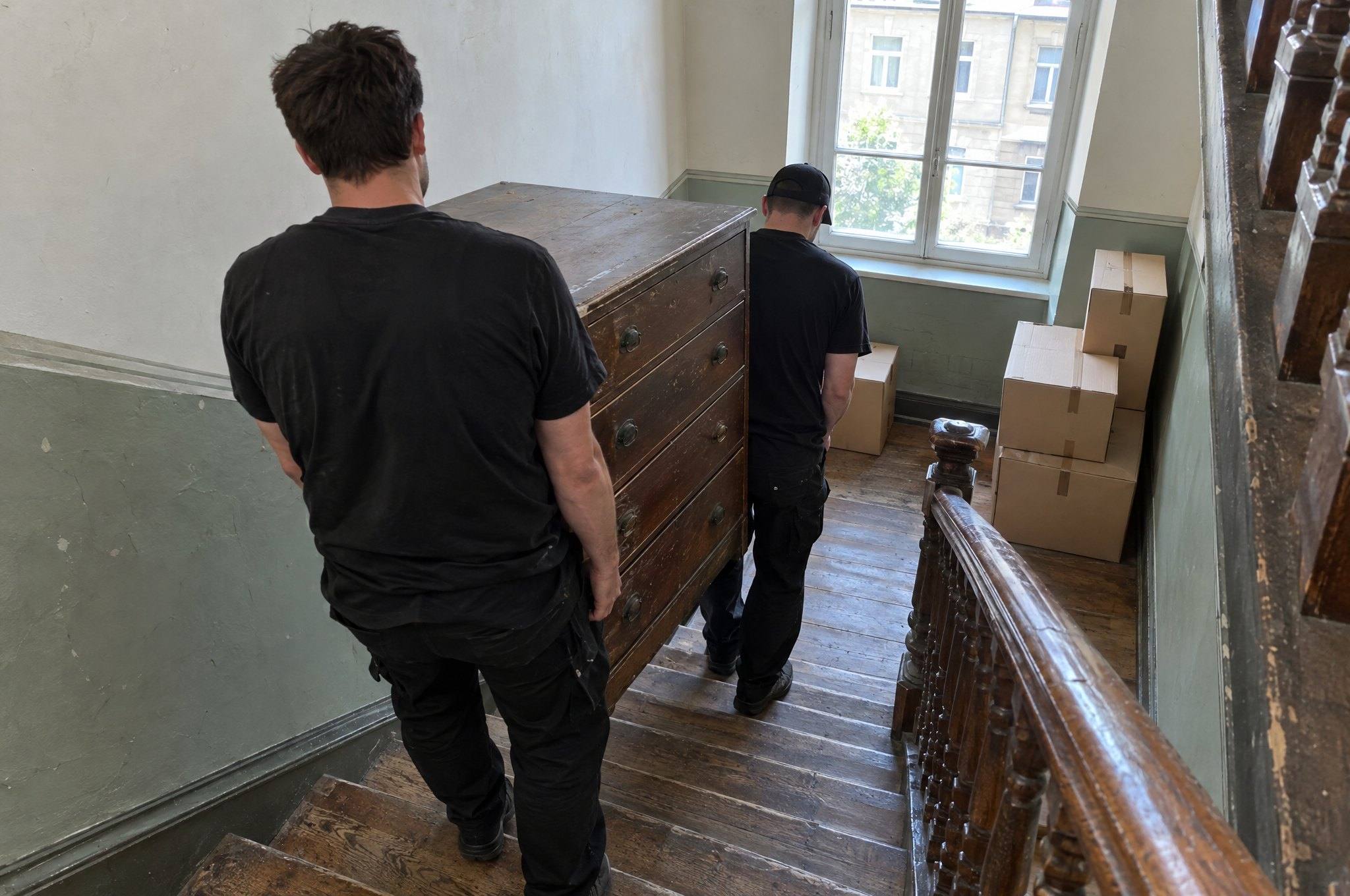Zwei Mitarbeiter tragen eine alte Kommode durch ein Altbau-Treppenhaus nach unten