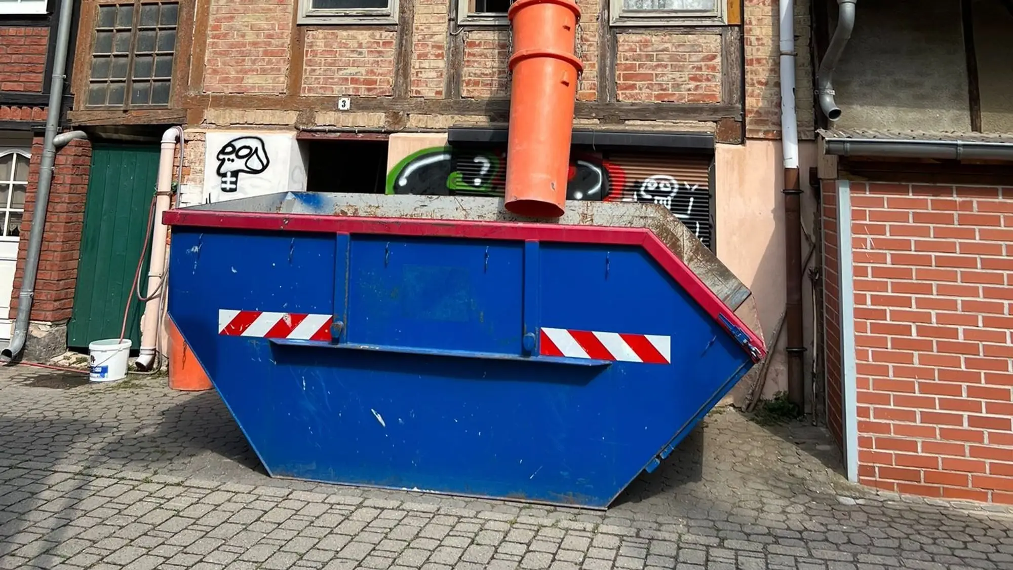 Blauer Absetzcontainer vor einem Fachwerkhaus, aus dem Fenster führt eine orange Schuttrutsche hinein