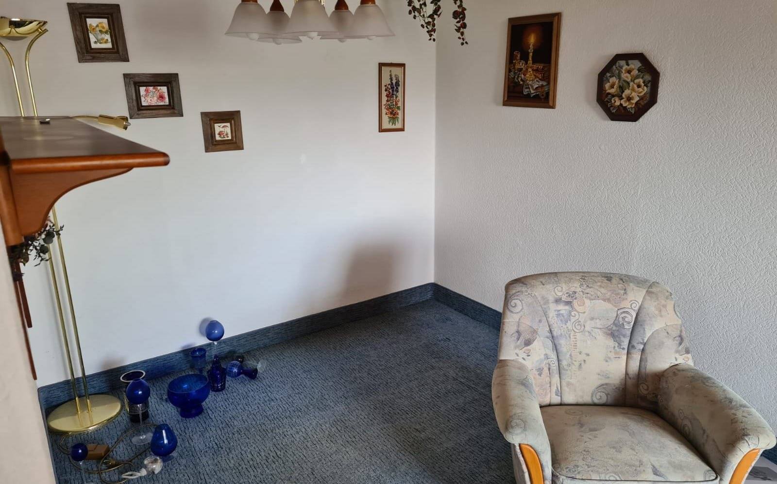 Wohnzimmer vor der Auflösung mit Sessel, Bildern an der Wand und Vasen auf dem Boden