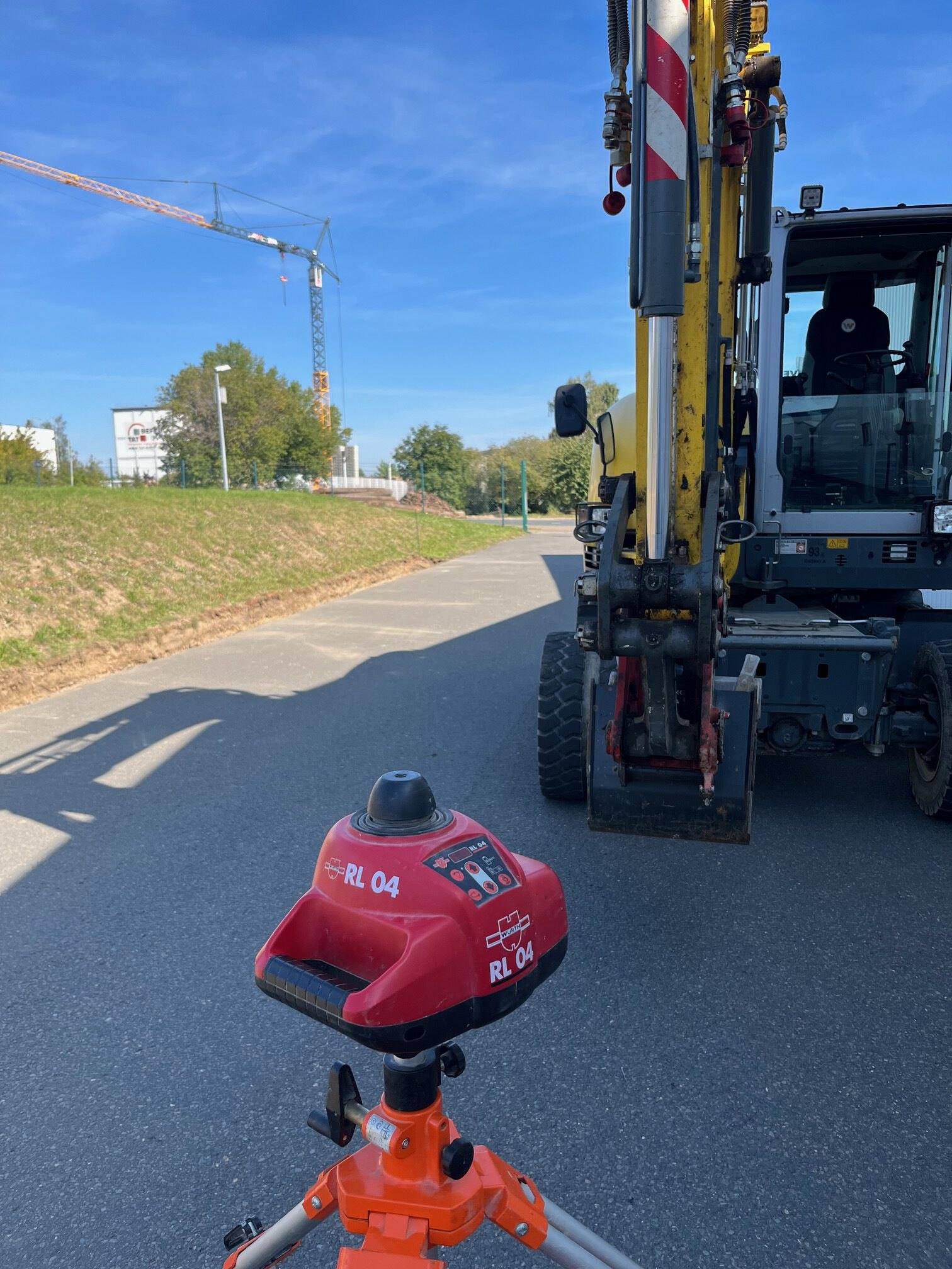 Rotationslaser auf Stativ vor einem Bagger an einer Straße